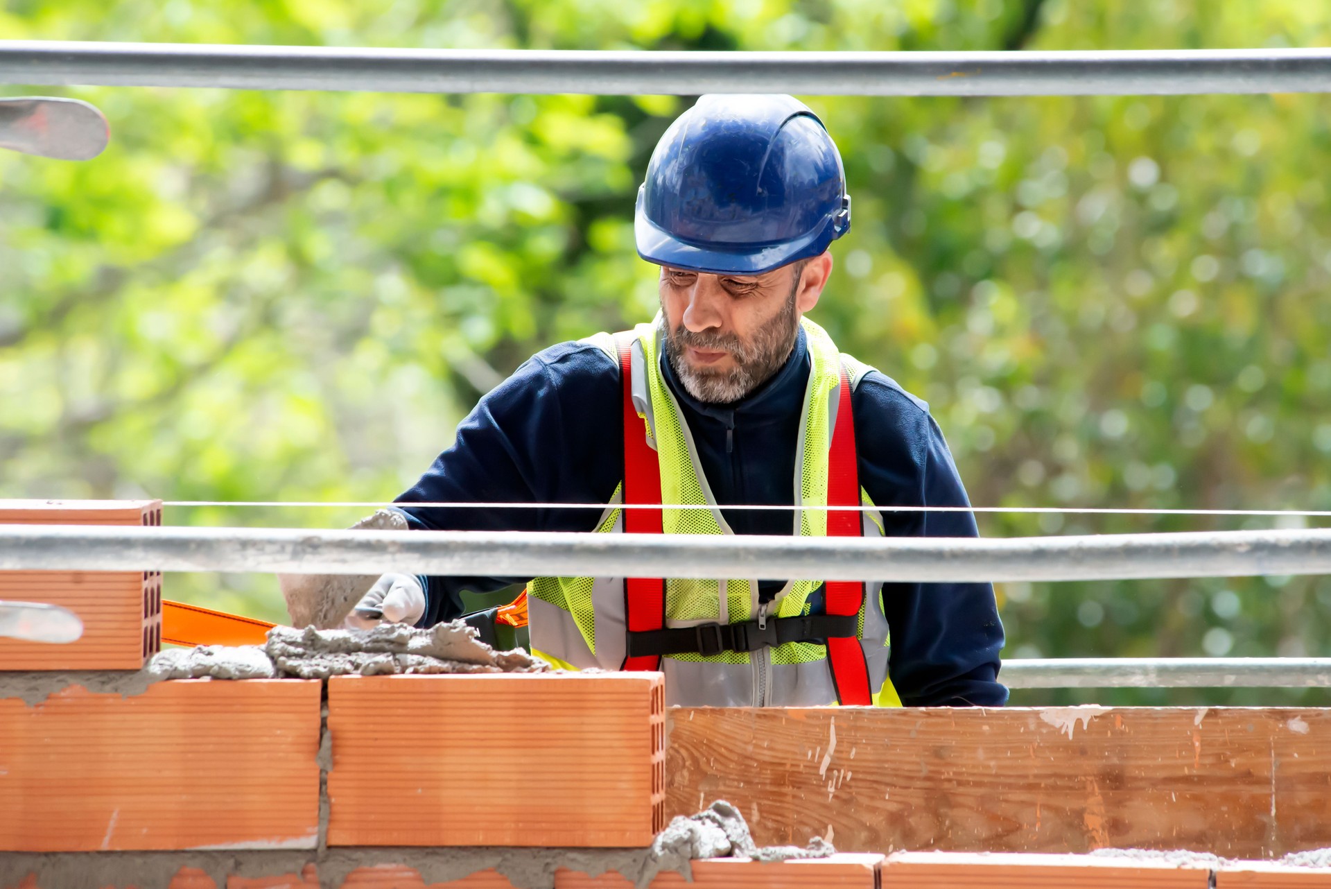 A master in a hard hat puts a cement mixture with a spatula on a brick against the backdrop of green trees at the height of a multi-story building under construction. The process brickwork of a wall.
